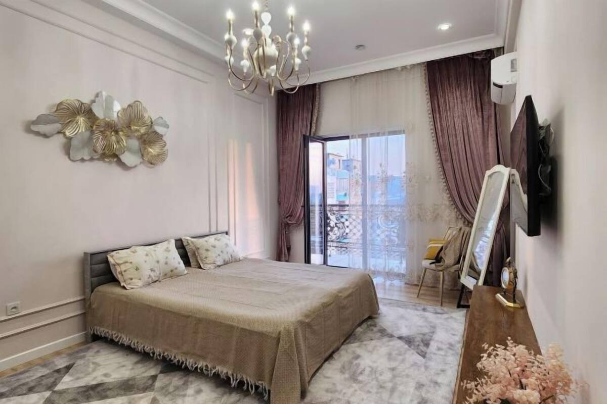 A bedroom with a bed, a chandelier, a large window with curtains, a mirror, and a dresser.