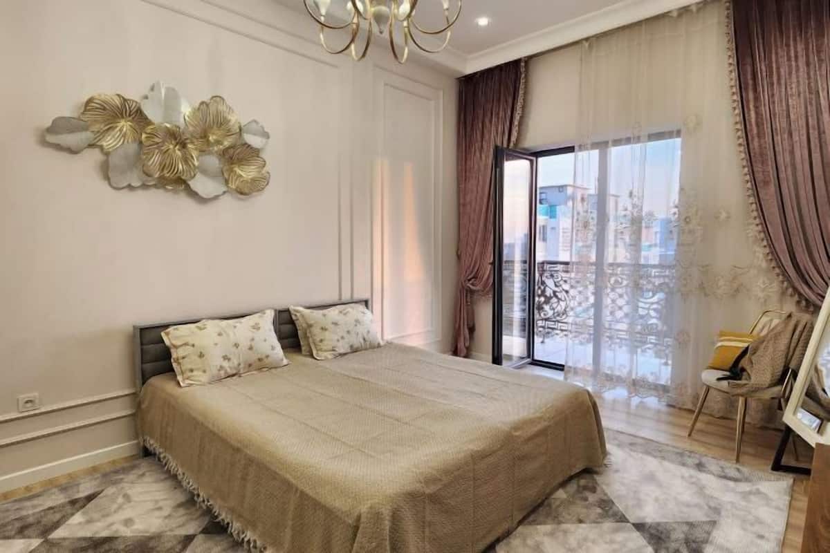 A bedroom with a large bed, a chandelier, a view through a glass door, and a chair by the window.