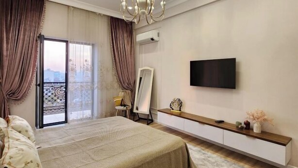 A bedroom with a large bed, a TV mounted on the wall, a vanity with a mirror, and a balcony with curtains. - Apartment Kazahstan in Tashkent (Tashkent)