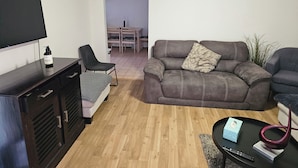 A living room with a sofa, a TV, a small table with a decorative item, and a wall clock. - Charming 3-bedroom House in Meadow Heights - Perfect Family Retreat (Melbourne)