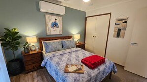 Un dormitorio con cama, mesitas de noche, ventilador de techo, aire acondicionado, una puerta y una obra de arte colgada en la pared.