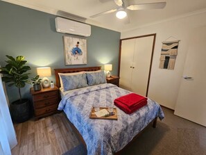 Un dormitorio con cama, mesitas de noche, ventilador de techo, aire acondicionado, una puerta y una obra de arte colgada en la pared.