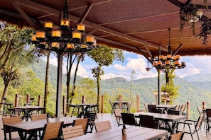 An outdoor dining area with tables and chairs, a wooden ceiling, and a view of mountains and trees.