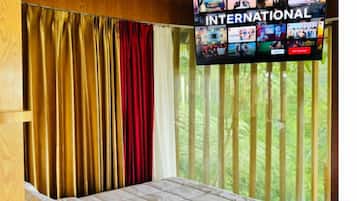 A hotel room with a bed, curtains, a television screen displaying various channels, and a window with vertical blinds.