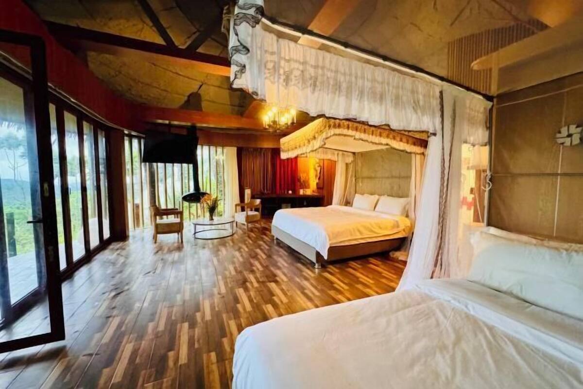 A spacious bedroom with a large bed, a bedside table, and a chair. There is a window with curtains and a view of the outdoors.