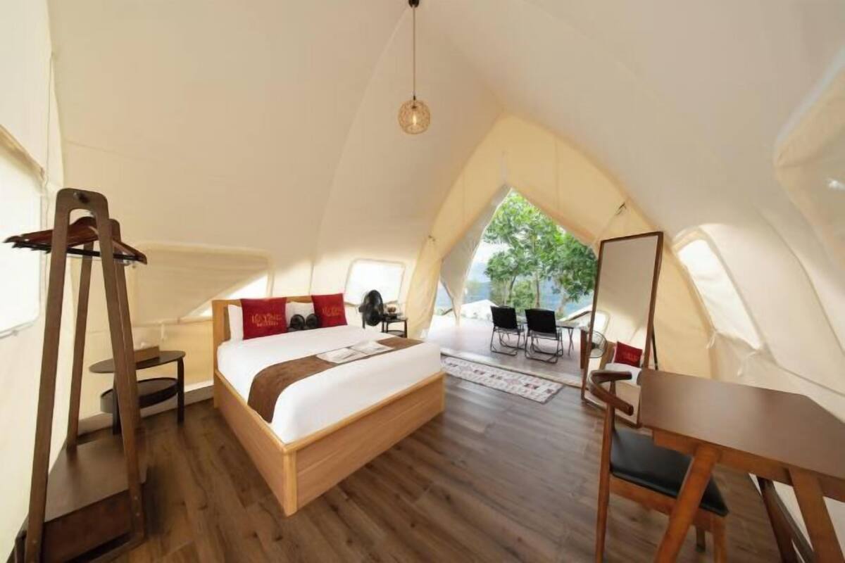 A bedroom with a wooden bed, a desk, and a chair, a small table, and a window with a view of greenery.