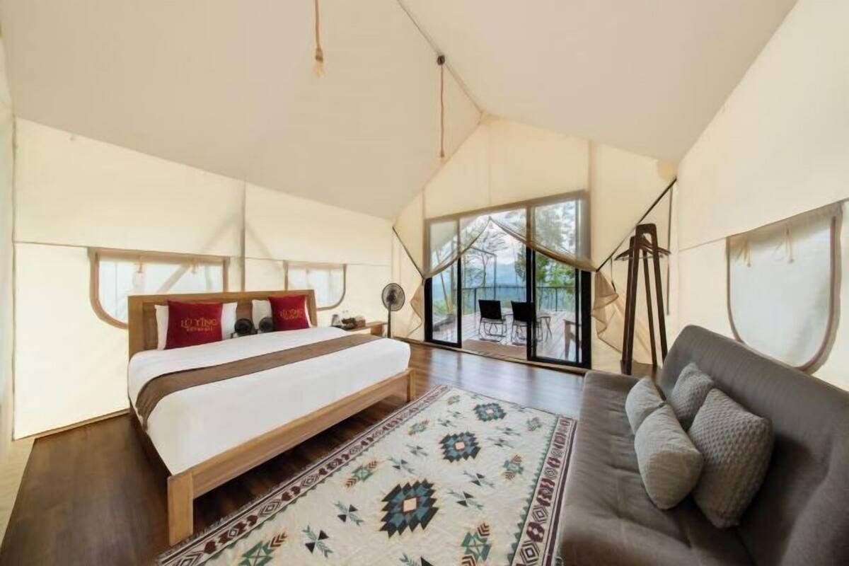 A bedroom with a large bed, a sofa, a patterned rug, and a view through large windows.