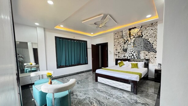 A modern bedroom with a large bed, a bench, and a seating area with a mirror and a vase of flowers. - Sathya Hotels & Resorts (Hyderabad)
