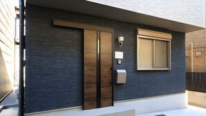A modern building with a dark brick facade, a large wooden door, and a small window with a light fixture. - Bliss Ikebukuro (Tokyo)