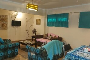 A living room with a sofa, armchairs, a coffee table, a TV, and a fan.