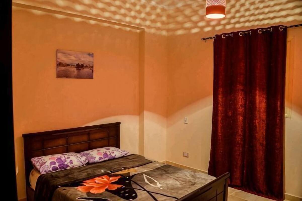 A bedroom with a bed, a window with curtains, a wall-mounted light fixture, and a small picture on the wall.