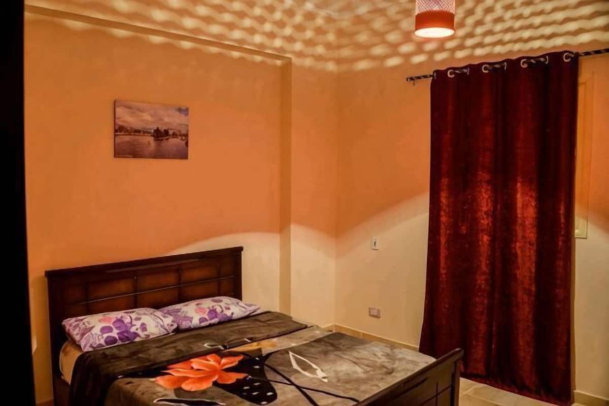 Ein Schlafzimmer mit Bett, einem fensterbelaubten Fenster, einer Wandleuchte und einem kleinen Bild an der Wand.