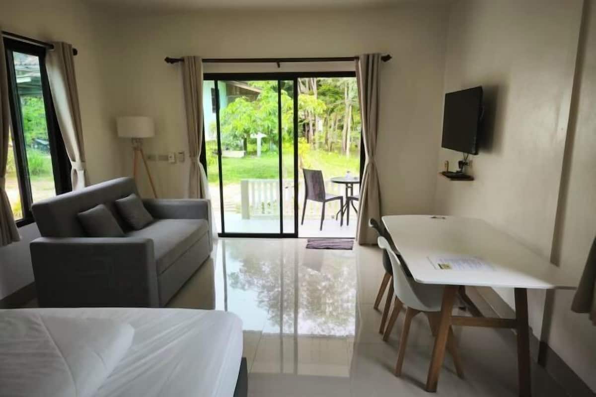 A hotel room with a bed, a sofa, a desk, a TV, and a sliding glass door leading to a garden.