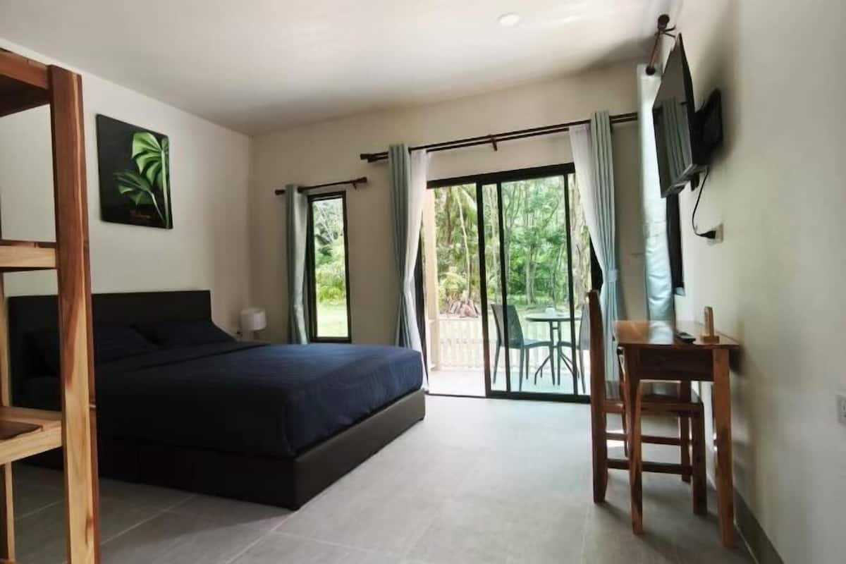 A bedroom with a bed, a desk, a chair, a TV, and a sliding glass door leading to an outdoor area.