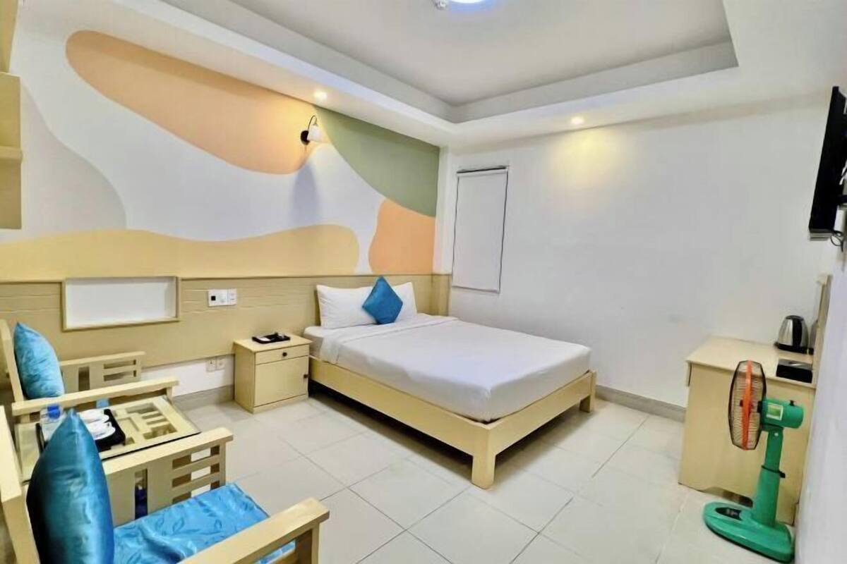 A hotel room with a bed, bedside tables, a desk, a chair, a TV, and a standing fan.