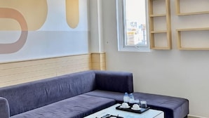 A modern hotel room with a gray sofa, a small table with a tray of water bottles and glasses, and a large abstract wall art piece. - Sea Charm Hotel (Da Nang)