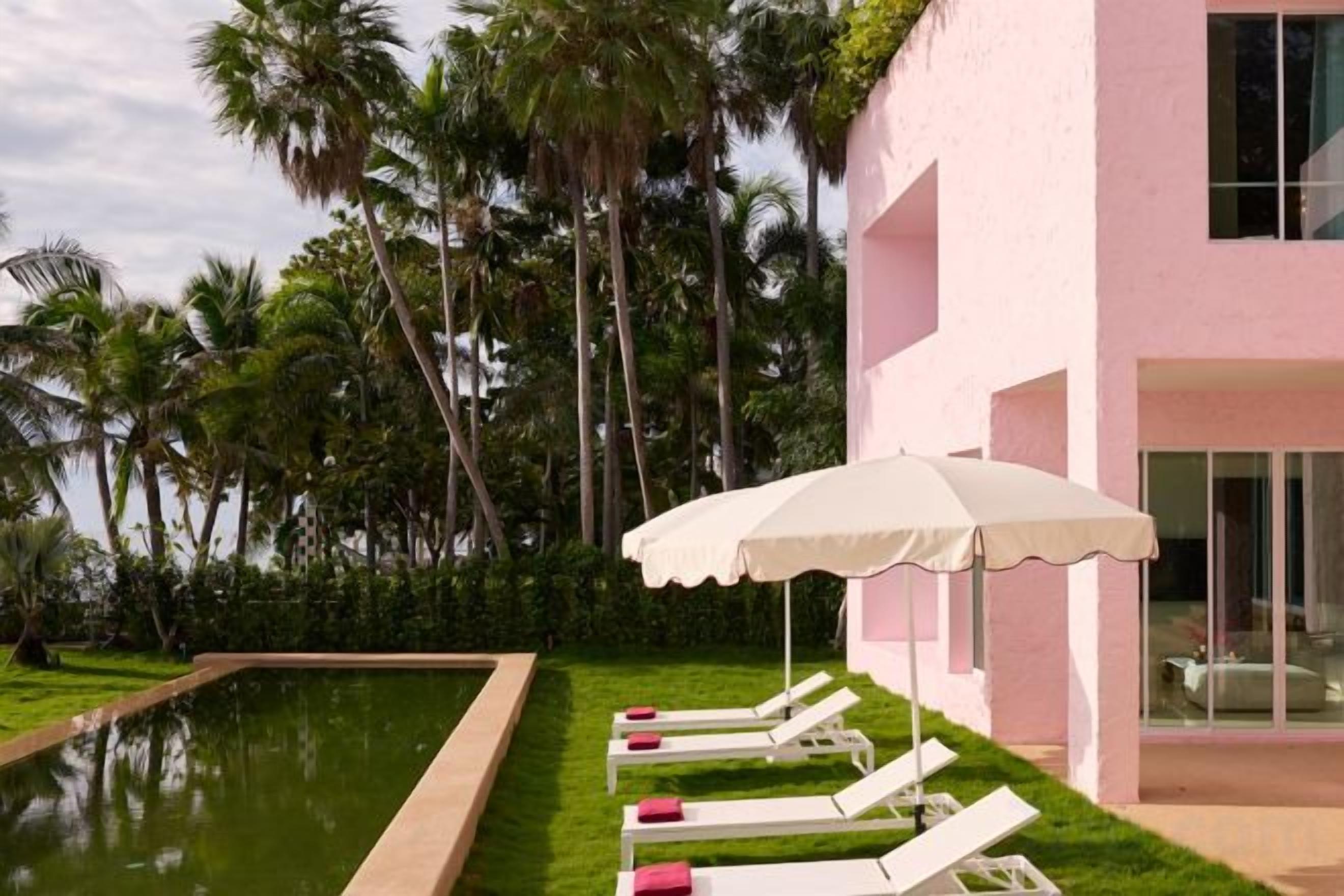 A pool area with lounge chairs and an umbrella, surrounded by palm trees and a pink building.