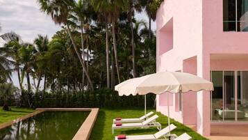 A pool area with lounge chairs and an umbrella, surrounded by palm trees and a pink building.