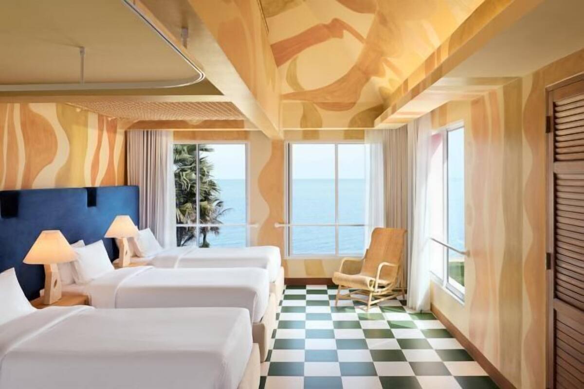 Quarto de hotel com duas camas, piso xadrez, vista para o mar e uma janela grande com cortinas.