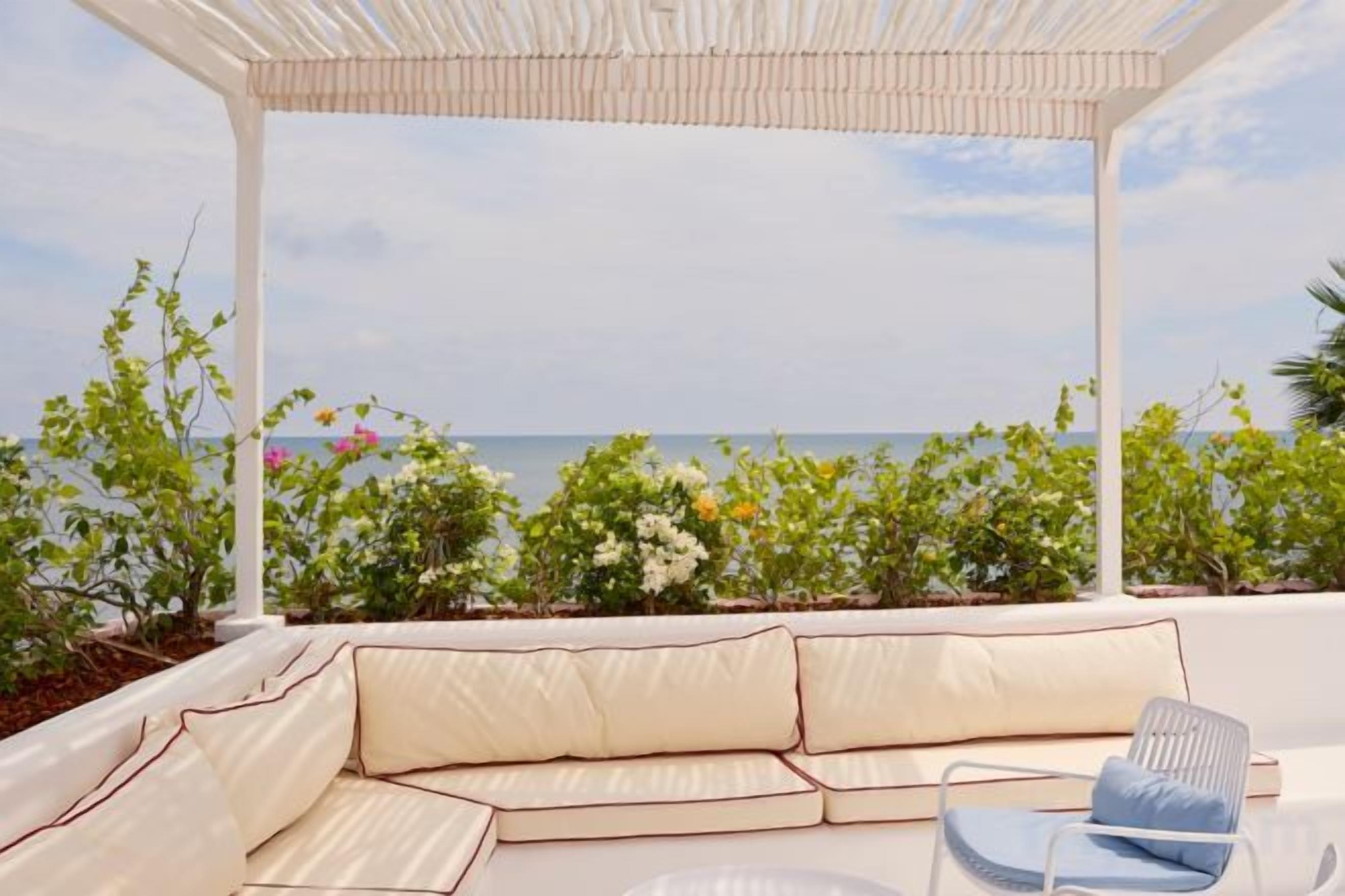 A covered outdoor seating area with a view of the sea, a cushioned sofa, and a chair.