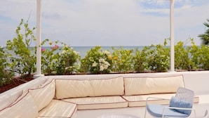 Un área de estar al aire libre techada con un sofá blanco acolchado y una sola silla, con vistas a un jardín lleno de plantas y flores, y vista al mar.