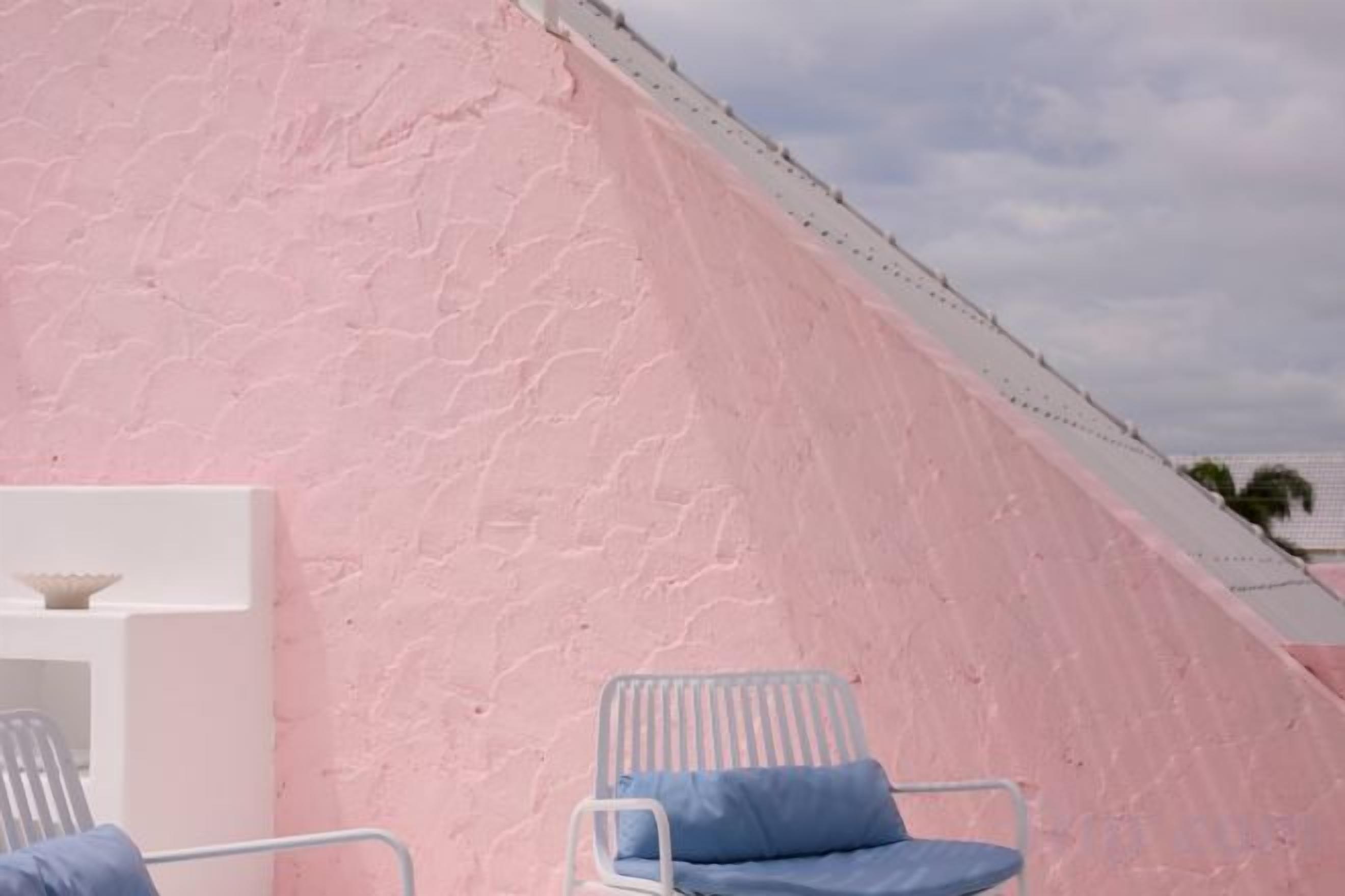 Pared rosa con textura, una repisa blanca y dos sillas blancas con cojines azules.