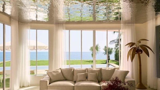 Un salón moderno con un sofá color crema, ventanas amplias con vista al mar y una lámpara de pie alta.