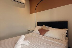 Un lit bien fait, avec du linge de lit blanc et une serviette pliée, une tête de lit noire et une petite table de chevet avec une lampe.