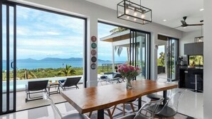 A modern dining area with a wooden table and chairs, large windows offering a view of the sea and mountains, and a kitchen area visible through the glass doors. - L'Esterel Villa - A Tropical Oasis at Maenam Hills (Koh Samui)