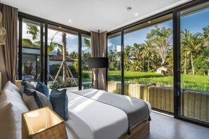 A modern bedroom with a large bed, a nightstand, a lamp, and a view of a lush garden through large windows. - Villa Juke by BaliSuperHost (Bali)
