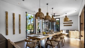 A modern dining area with a long wooden table, multiple chairs, and hanging pendant lights.