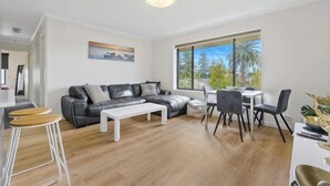 A modern living room with a sectional sofa, a dining table, and chairs. There is a large window providing a view of the outdoors. - Beachside Breakaway Glenelg South – 200m to the Beach (Glenelg South)