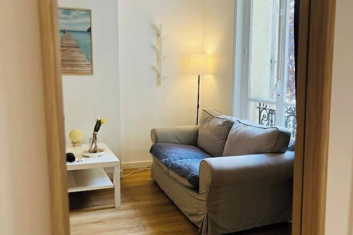 Ein Zimmer mit einem beigen Sofa, einem kleinen weißen Tisch mit Lampe und einem Gemälde eines Piers.