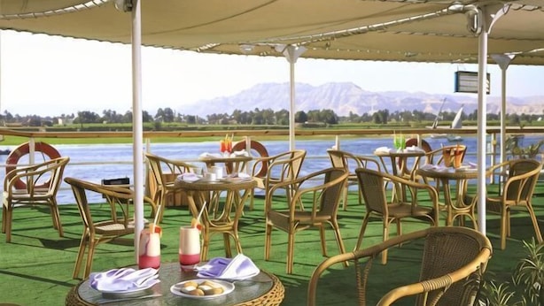 An outdoor seating area with wicker chairs and tables, a canopy, and a view of a lake and mountains. - Jaz Crown Prince Nile Cruise - Every Monday from Luxor for 07 & 04 Nights (At Tod)