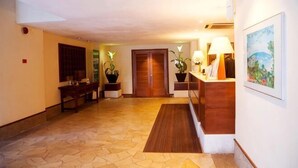 En hotellobby med en receptionsdisk, trädörrar, krukväxter, en tavla och ett mönstrat golv.