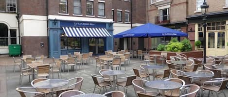 An outdoor seating area with tables and chairs, a building with a blue awning, and a brick facade.