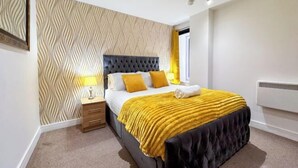 Une chambre à coucher avec une tête de lit capitonnée, un lit recouvert d’une couverture jaune, une lampe de chevet et un papier peint à motifs.