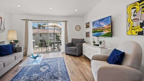 A modern living room with a sofa, armchair, coffee table, and a view through a glass door to an outdoor patio. - Palm Springs Condo with Pool & Mt Views Sleeps 4 (Palm Springs)