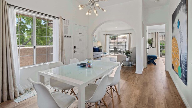 A modern dining area with a glass table and white chairs, a chandelier, and a view of the outdoors through large windows. - Palm Springs Condo with Pool & Mt Views Sleeps 4 (Palm Springs)