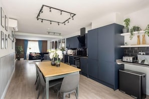 A modern kitchen with dark cabinetry, a dining area with a wooden table and chairs, and a living room with a sofa and curtains.