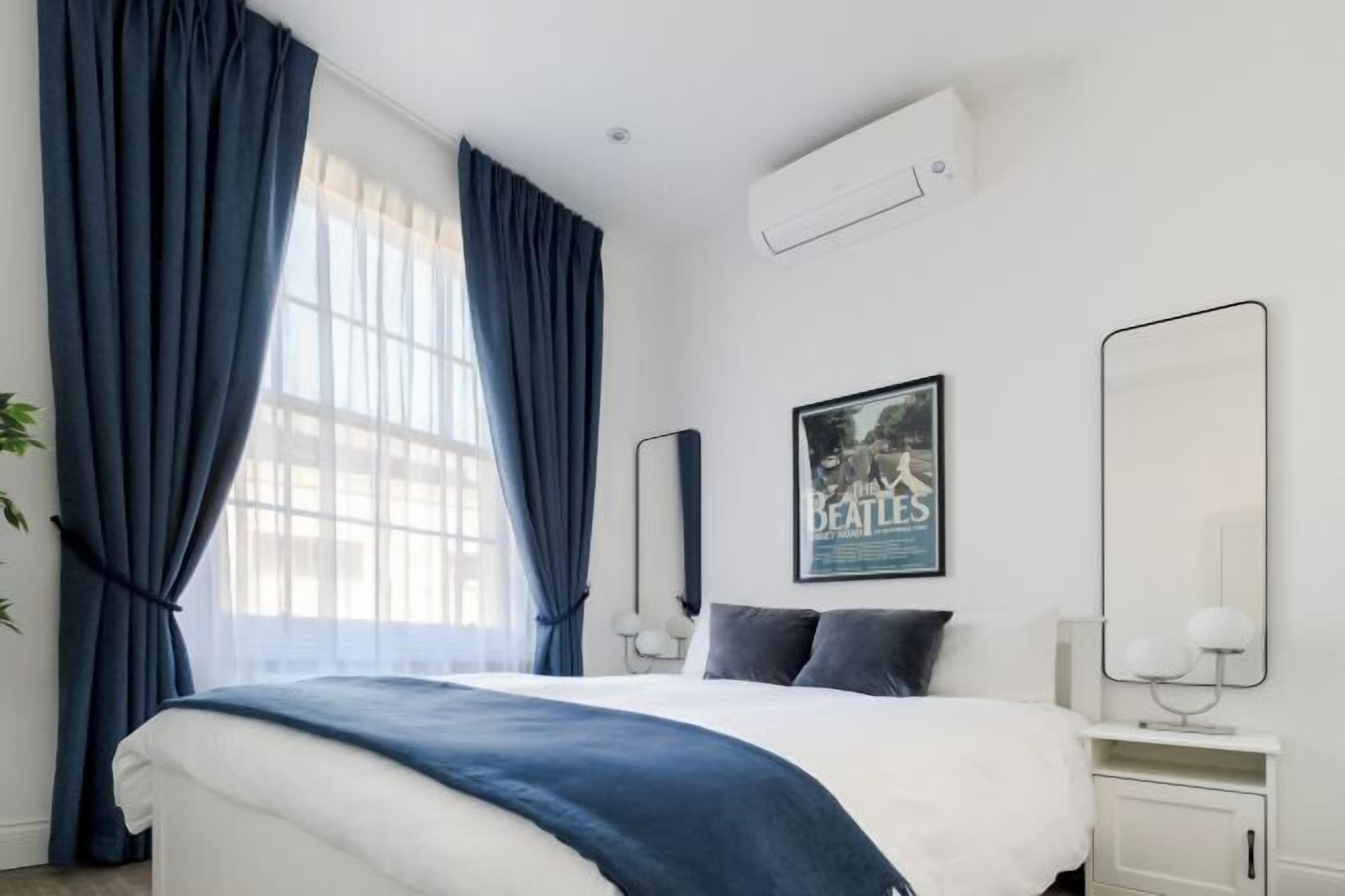 A modern hotel room with a large bed, a window with curtains, a poster on the wall, and a mirror.