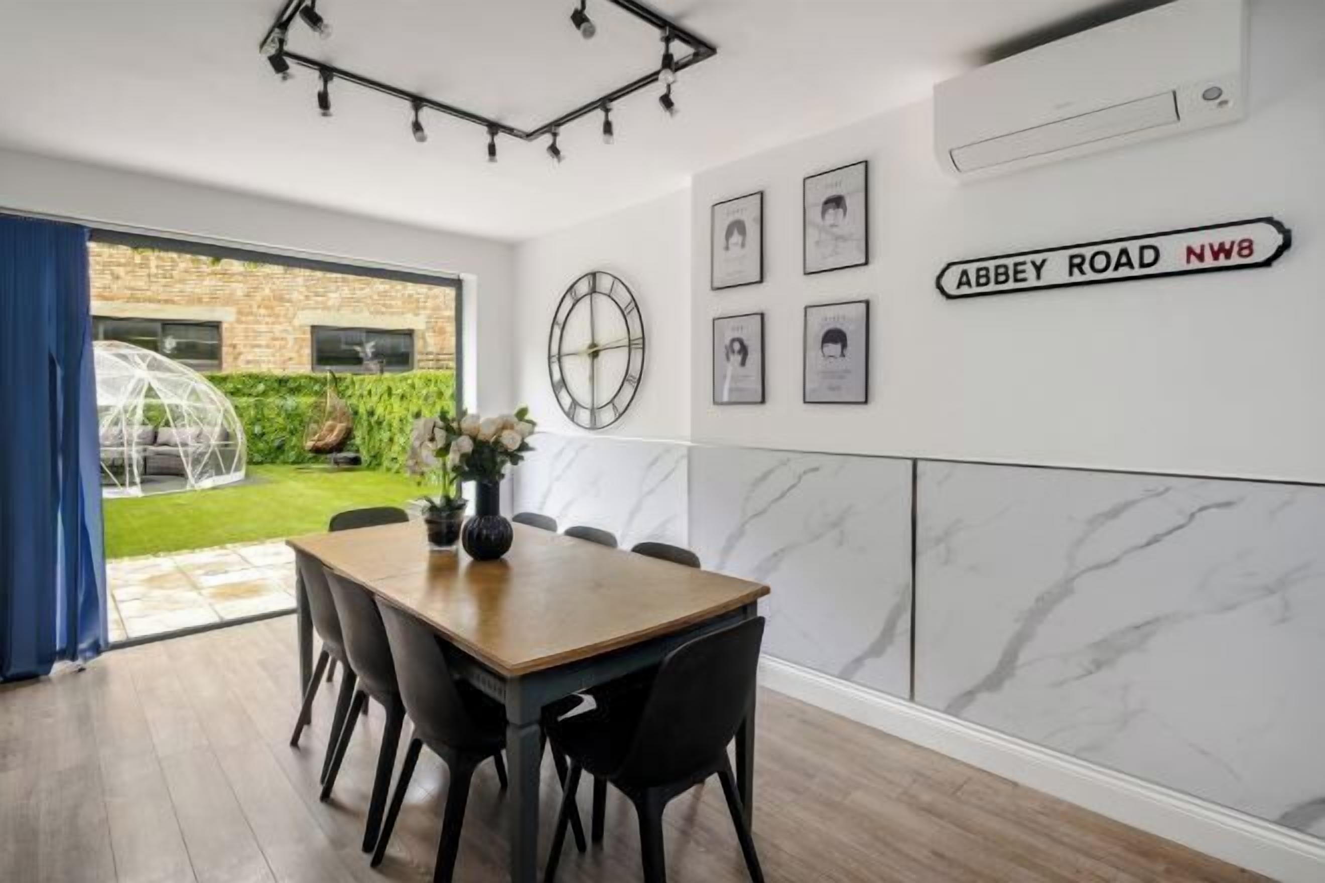 A dining area with a wooden table and chairs, a large window with a view of a garden, and a wall clock.