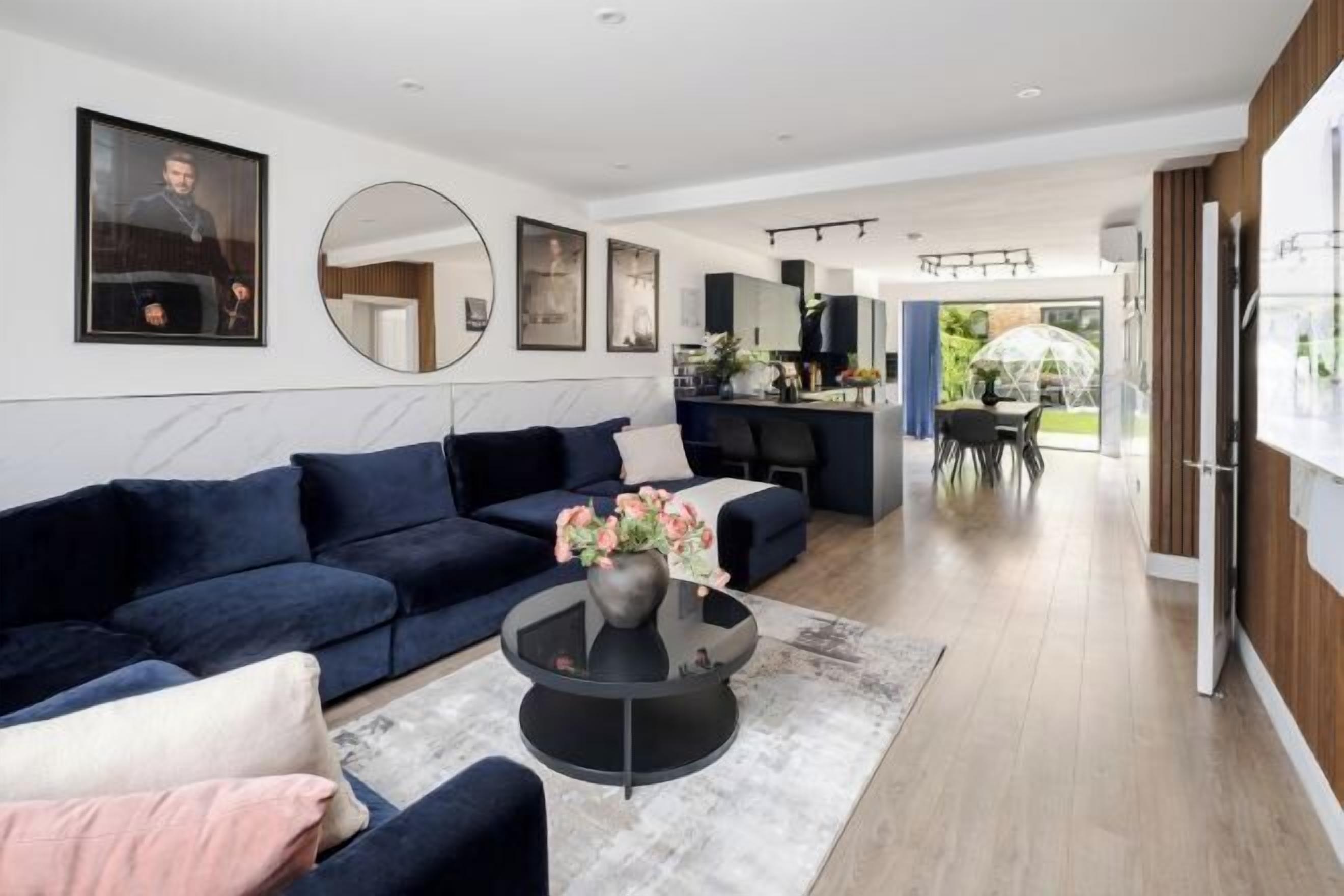 A modern living room with a dark blue sofa, a round coffee table with flowers, and a kitchen in the background.