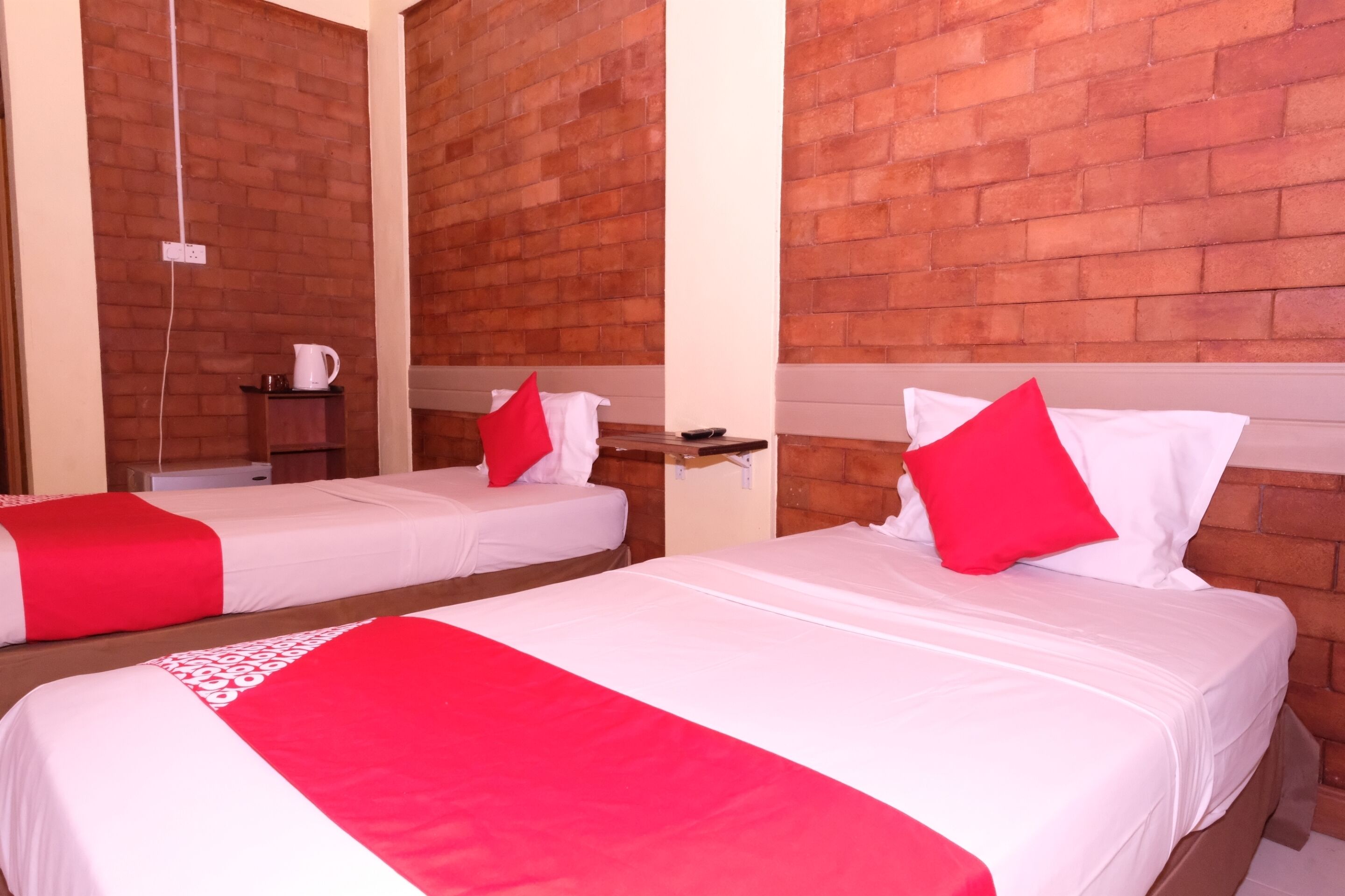 Dos camas individuales con ropa de cama roja y blanca, una mesita con una tetera y una pared de ladrillo.