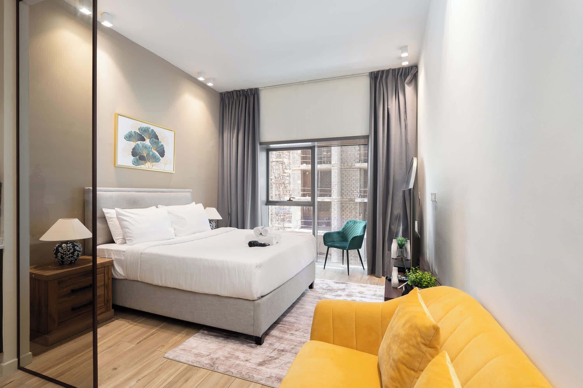 A modern bedroom with a large bed, a sofa, a desk, and a window with a view of the city.