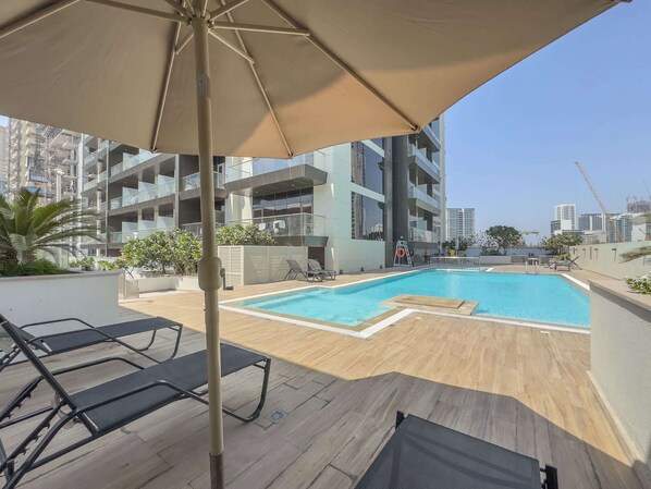 A rooftop swimming pool with lounge chairs and an umbrella, surrounded by modern buildings and a clear sky.