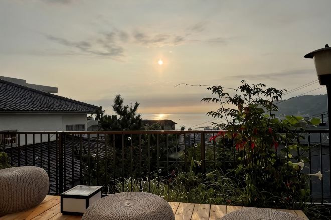 A balcony with a view of the sunset over the sea, surrounded by greenery and a fence.