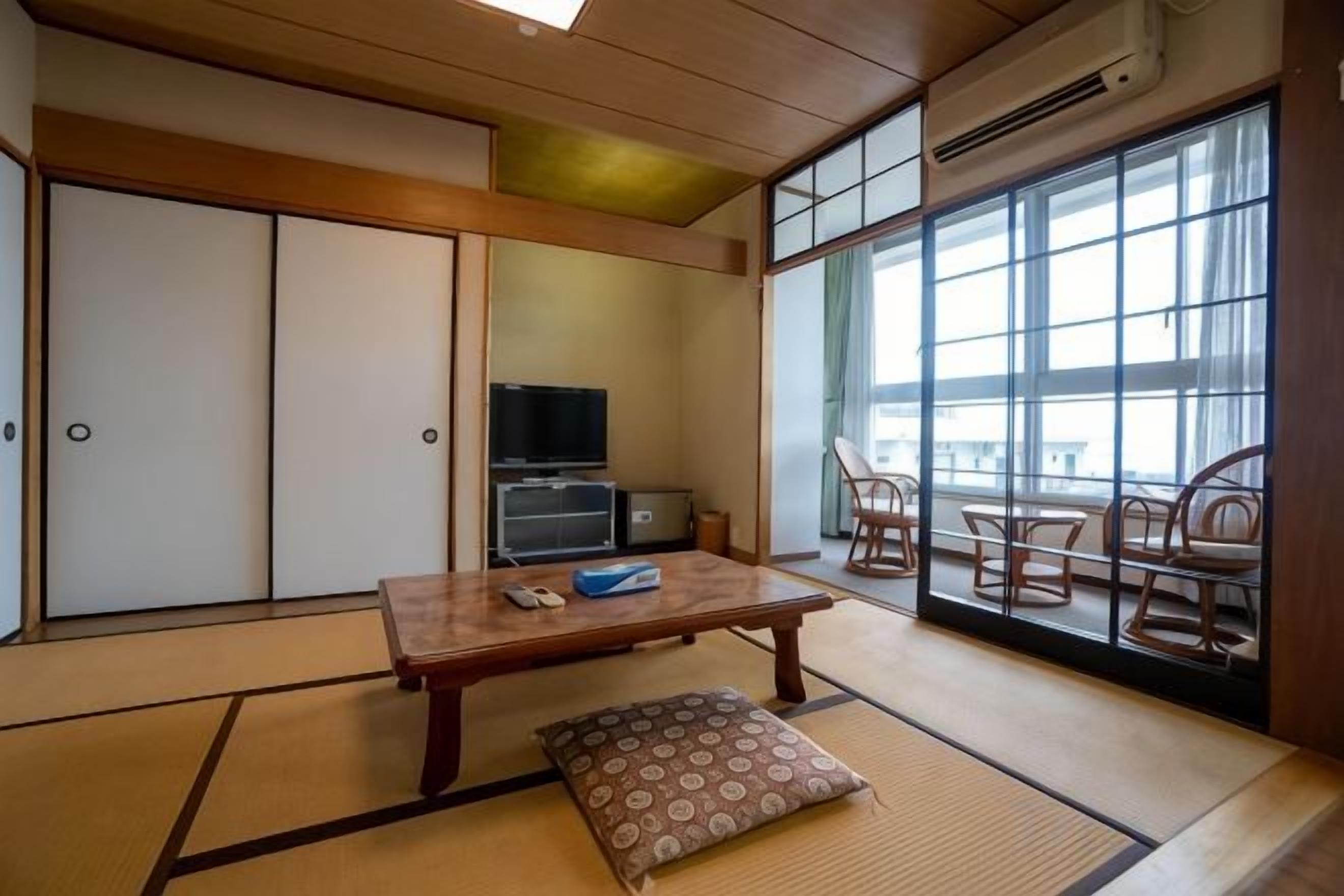 Una habitación japonesa tradicional con suelo de tatami, una mesa baja, un cojín, un televisor y puertas correderas.