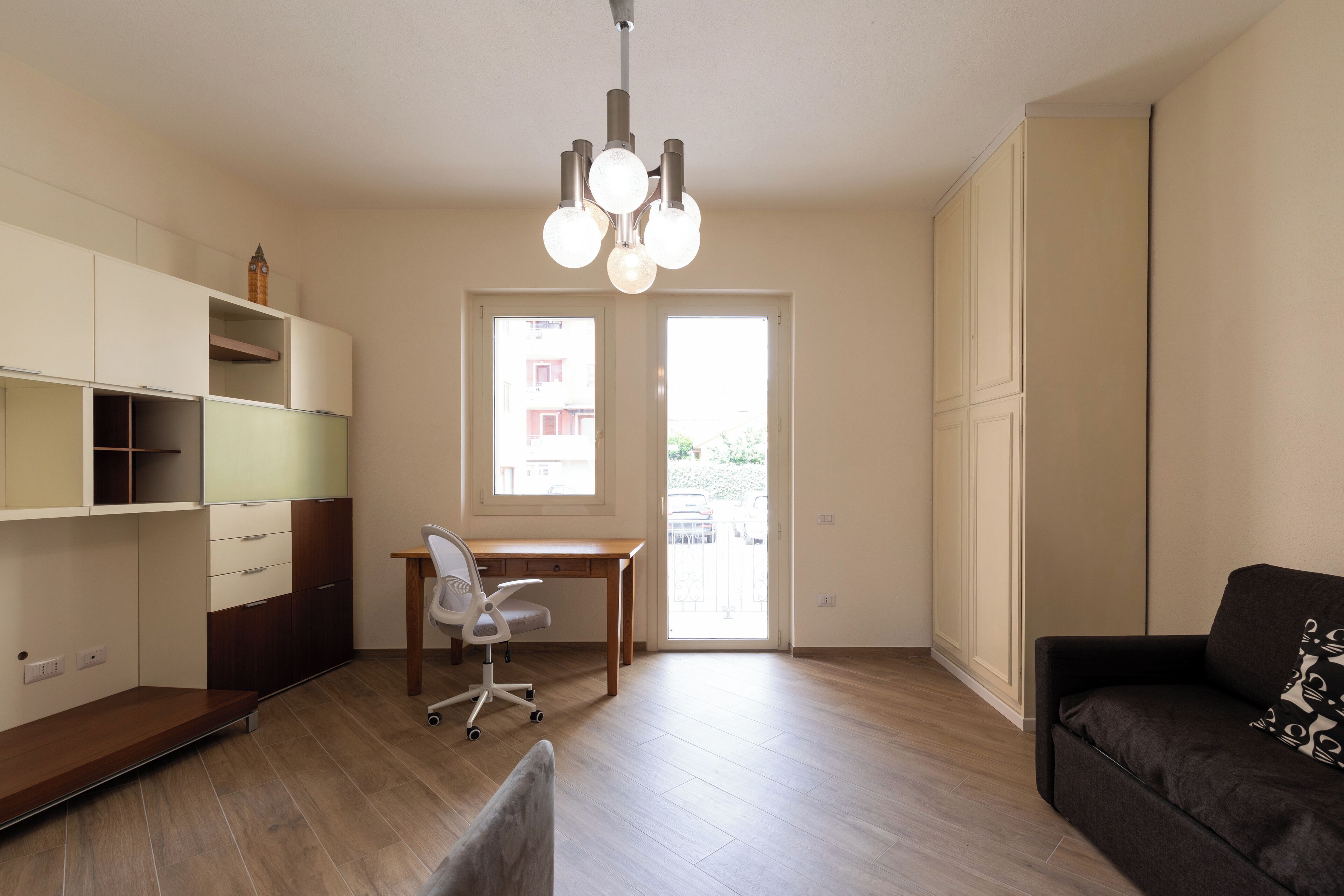 A modern living room with a wooden floor, a desk with a chair, a sofa, a built-in wardrobe, and a chandelier.