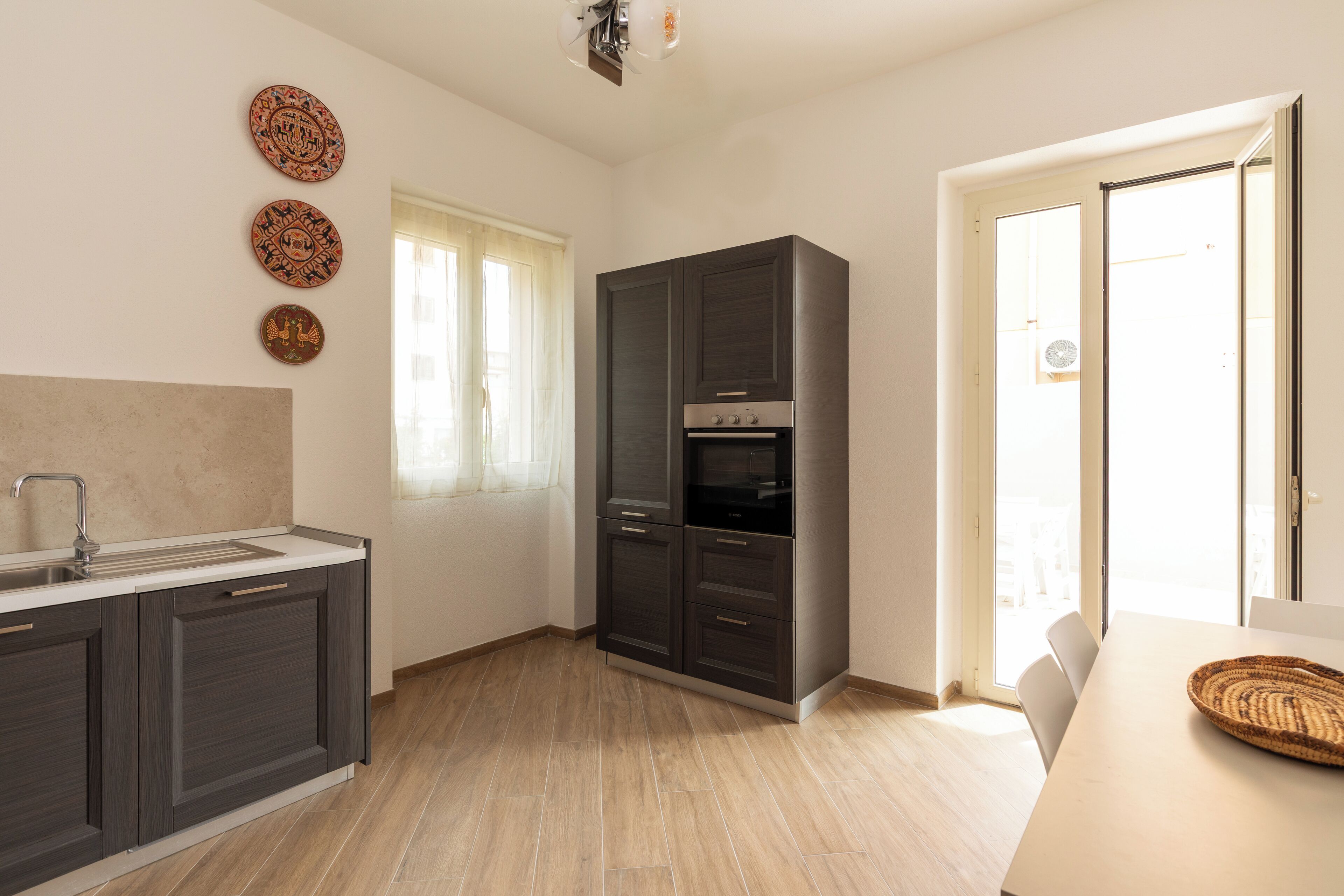 A modern kitchen with dark cabinets, a built-in oven, and a window with sheer curtains.